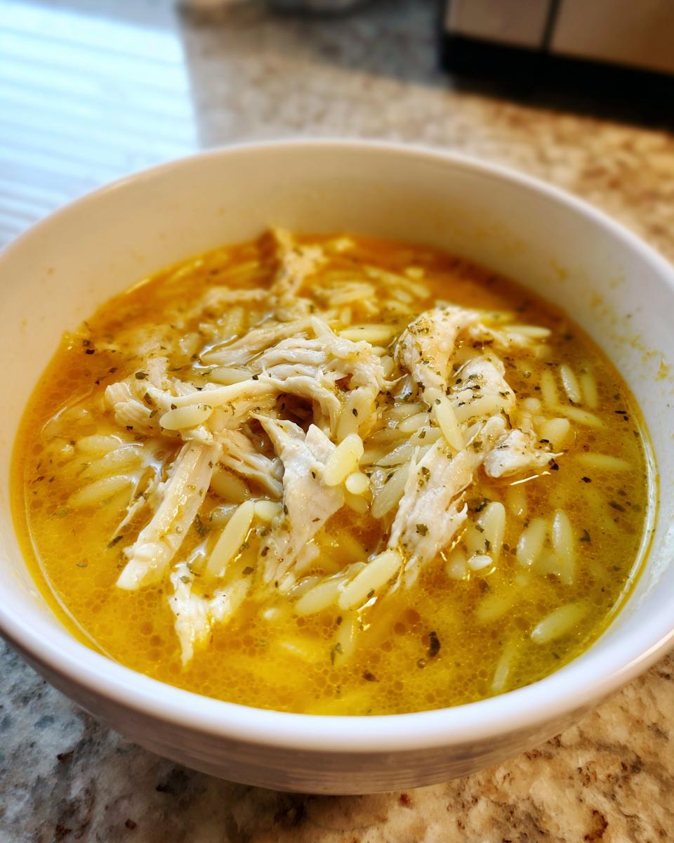 Close-up of a white bowl filled with rich, yellow chicken orzo soup topped with shredded chicken and herbs.
