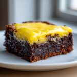 Close-up of a fudgy, dark chocolate brownie square topped with a thick, bright yellow lemon layer, served on a white plate.