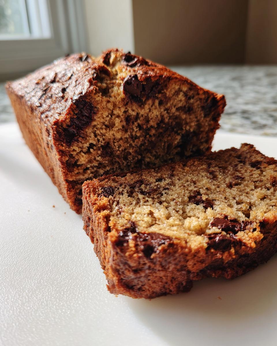 A close-up of a moist chocolate chip banana bread loaf with one slice cut and resting beside it.