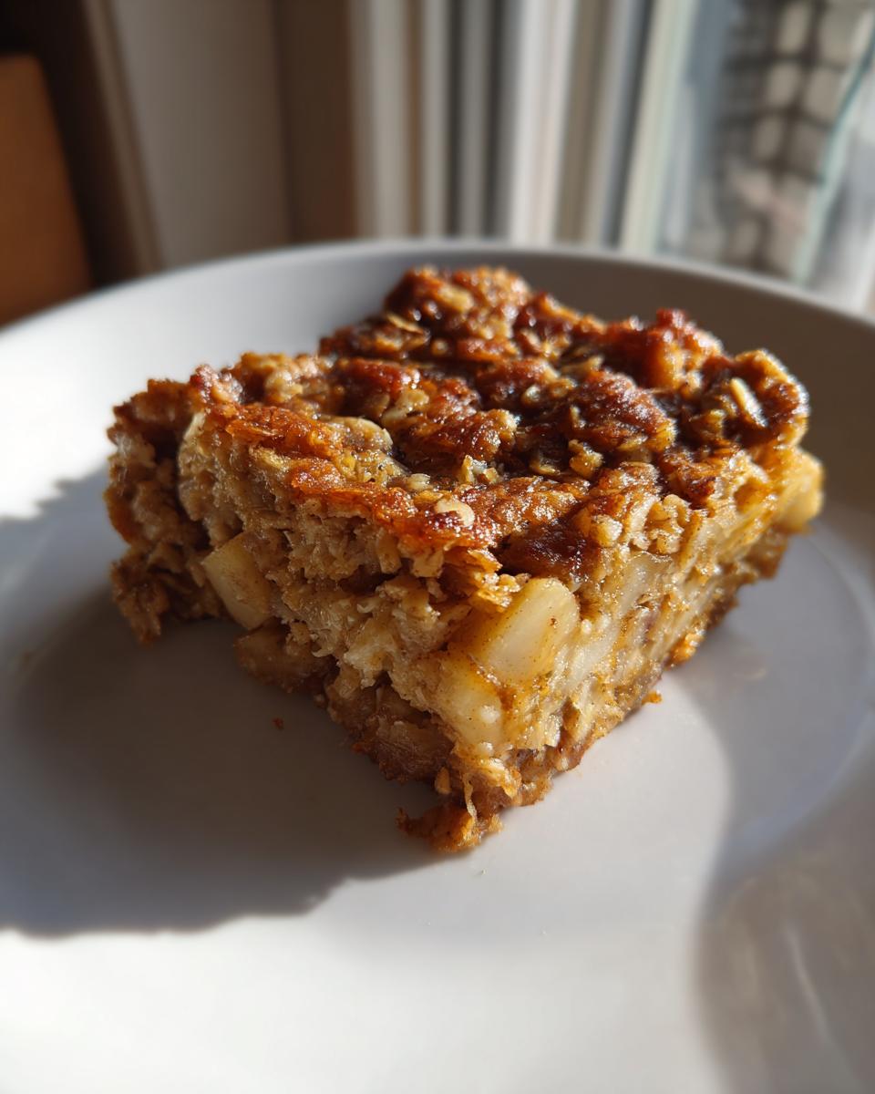 A close-up of a square slice of baked oatmeal with visible apple chunks, served on a white plate.