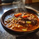 A steaming, dark brown bowl filled with rich beef barley soup, showing chunks of beef, barley, carrots, and celery.
