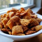 A close-up shot of a white bowl filled with golden brown, crunchy pieces from the toffee chex mix recipe.