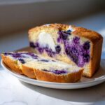 A loaf of Blueberry Cream Cheese Bread sliced on a white plate, showing a rich swirl of purple blueberries and cream cheese filling.