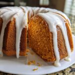 Close-up of a moist, orange sweet potato pound cake with thick white glaze dripping down the sides, one slice removed.