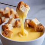 A piece of bread is lifted from a white bowl of melted cheese fondue recipe, showing a long, stretchy cheese pull.