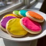 A plate of round sugar cookies topped with vibrant, neon-colored cookie icing in shades of pink, yellow, and purple.